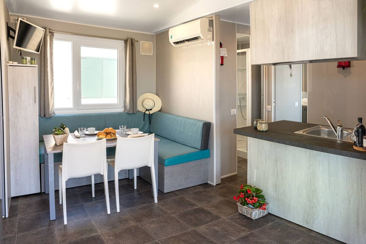 Ett litet, praktiskt kök med en diskho och en arbetsbänk, en matplats med ett bord och stolar, en blå, vadderad bänk och ett fönster med gardiner.