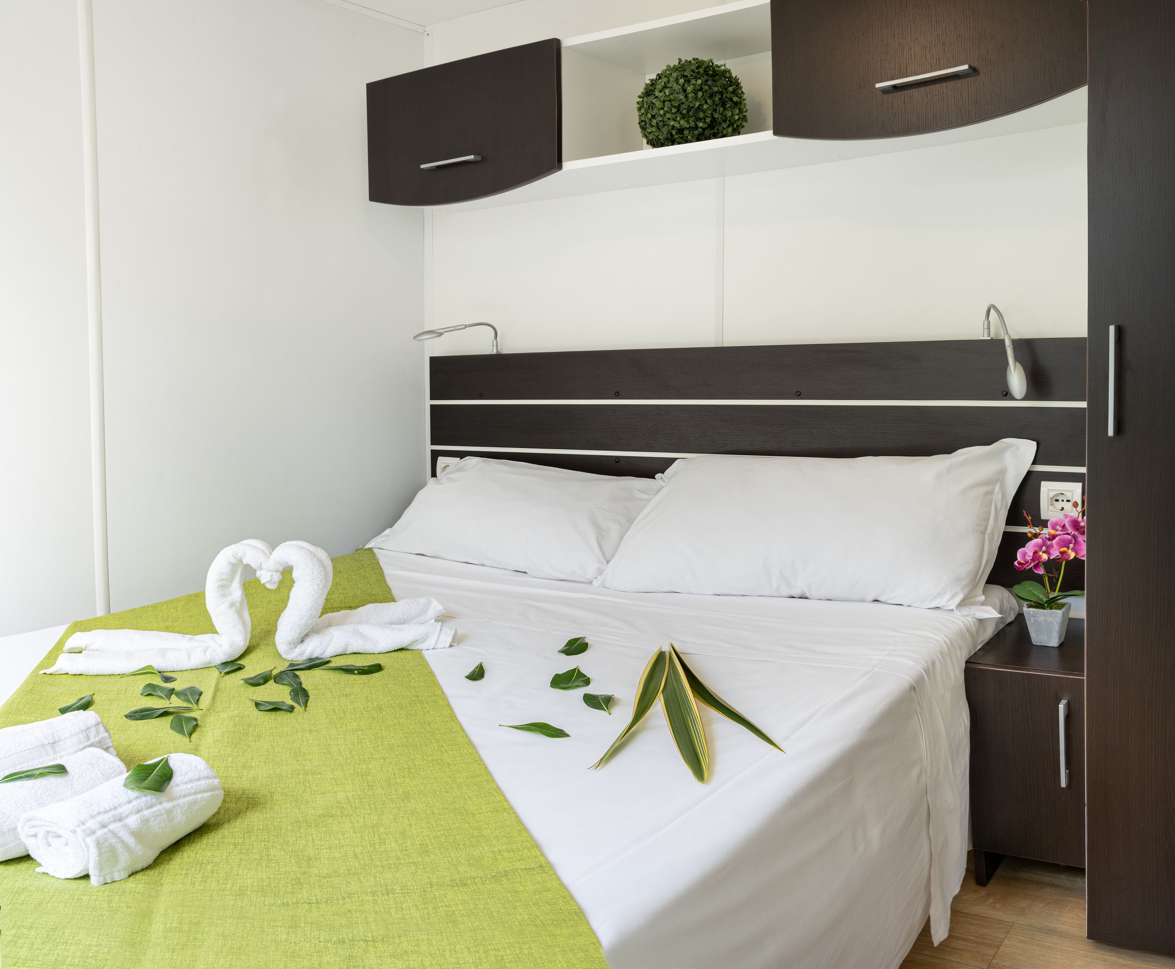Ein ordentlich bezogenes Bett mit weißen Leinentüchern, ein grünes Handtuch und eine Anordnung von Handtüchern in Herzform. Grüne Blätter sind auf dem Bett verteilt, und auf dem Nachttisch steht ein kleiner Blumentopf mit rosa Blüten.
