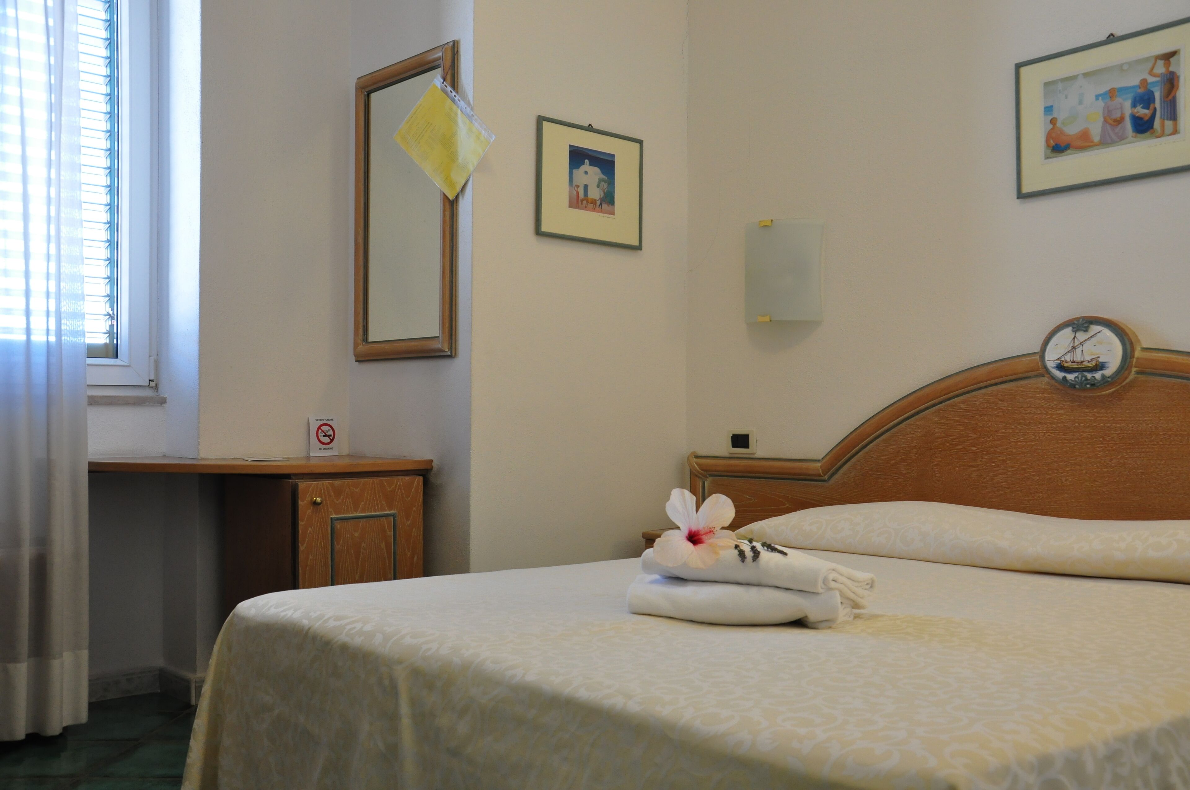 Quarto de hotel com cama, cabeceira de madeira, mesa de cabeceira, espelho e relógio fixado na parede.