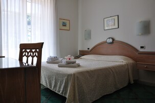 Una cama bien tendida con cabecera de madera, una silla de madera, una mesita y una ventana con cortinas traslúcidas.