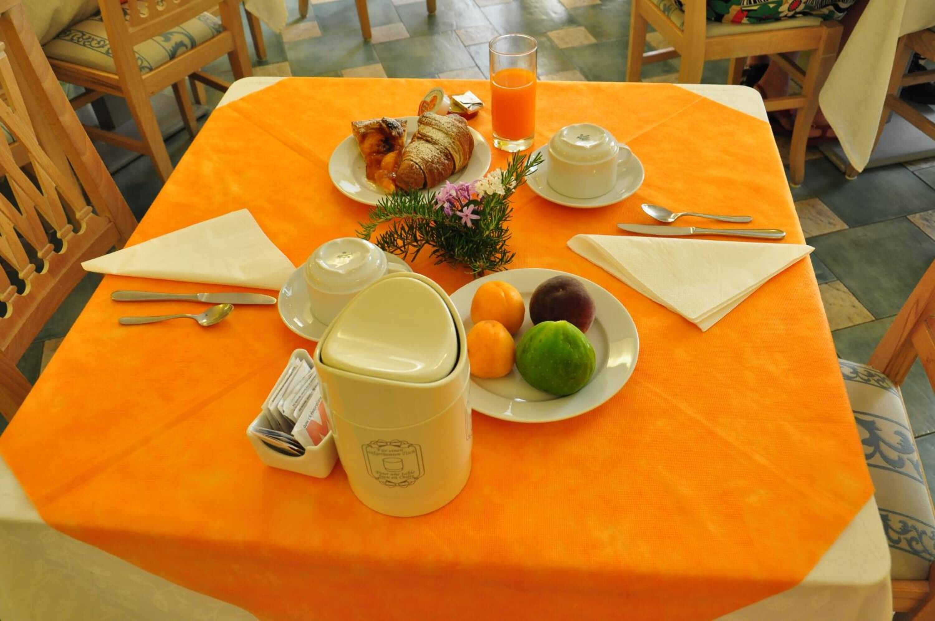 Ein Frühstückstisch mit einem Croissant, Orangensaft, einer Obstplatte, einer Kaffeekanne und einer Zeitung.