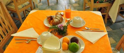 Ein Frühstückstisch mit einem Croissant, Orangensaft, einer Obstplatte, einer Kaffeekanne und einer Zeitung.