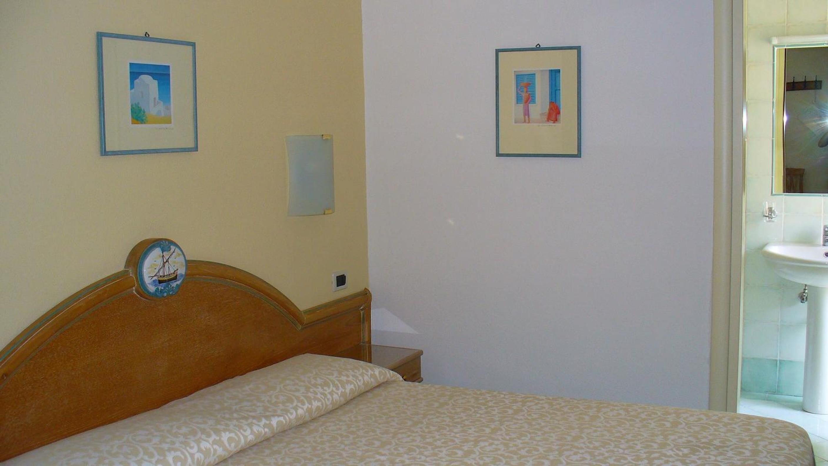 Habitación de hotel con cama, un reloj de pared, dos cuadros enmarcados, una mesita de noche y un baño con lavamanos y espejo.