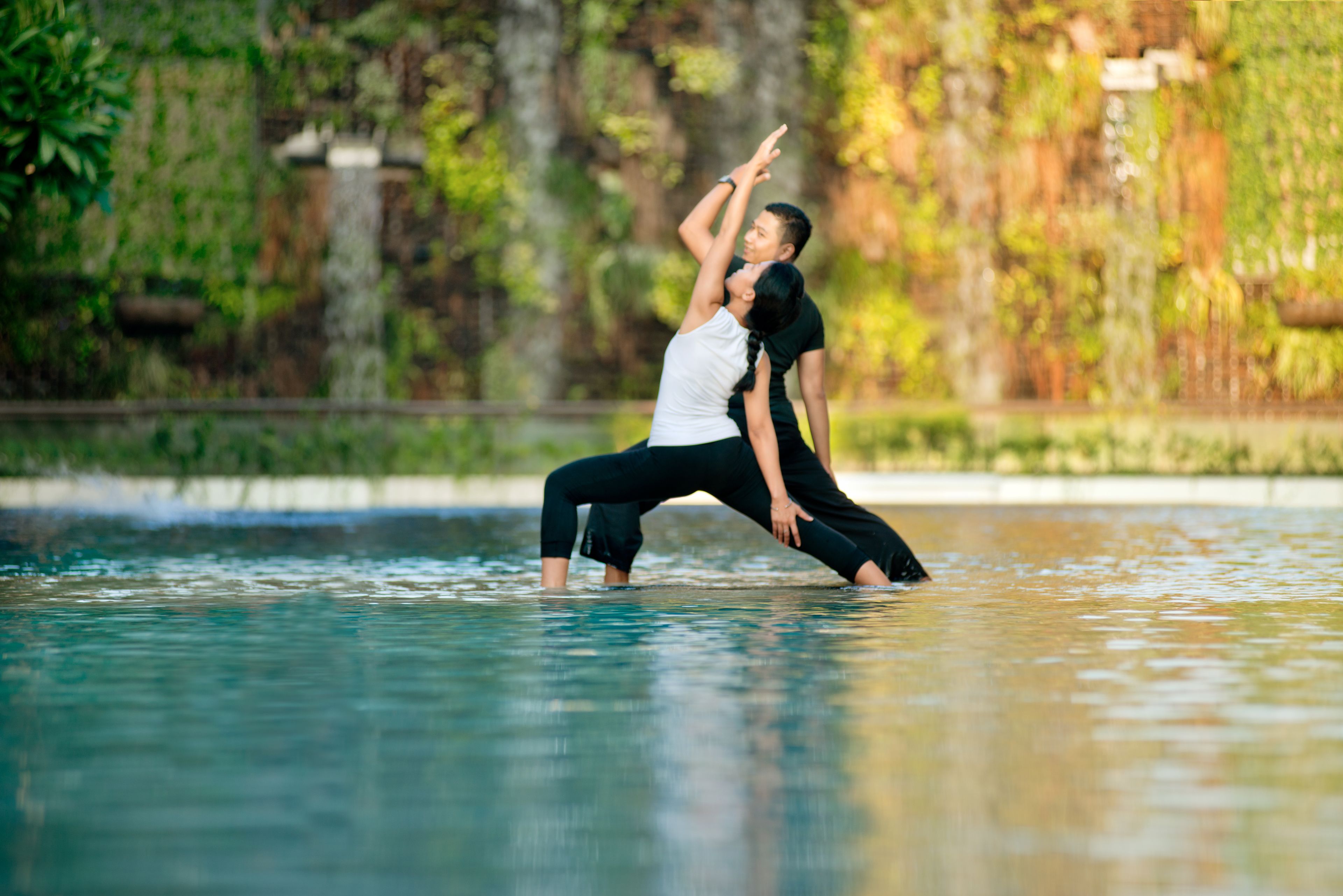 Two people performing a yoga pose in a water body surrounded by greenery.
