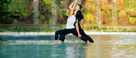Two people performing a yoga pose in a water body surrounded by greenery.