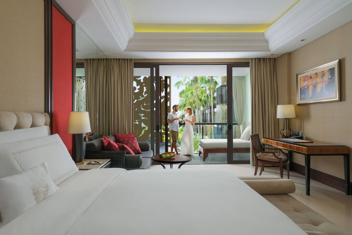 A hotel room with a large bed, a desk, a chair, and a sofa. There is a view of palm trees and a balcony with a person standing.
