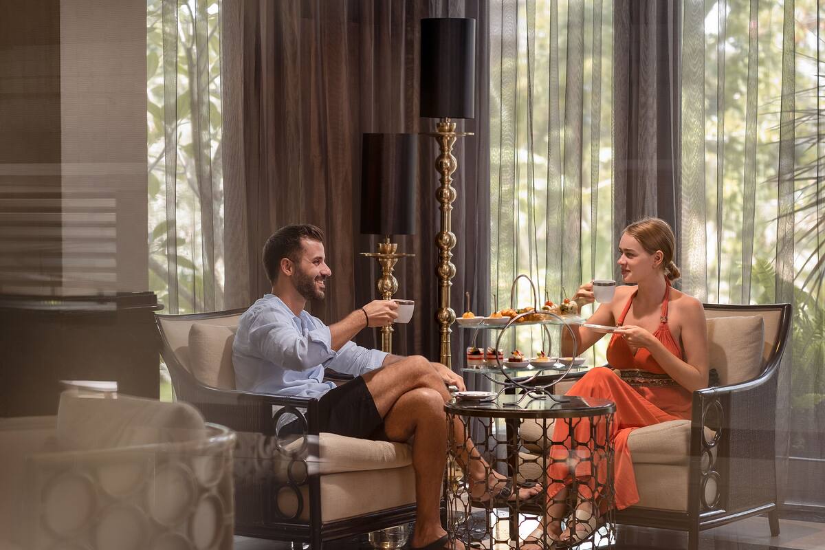 A couple seated on a sofa in a room with a large window, a table with food, and a decorative lamp.