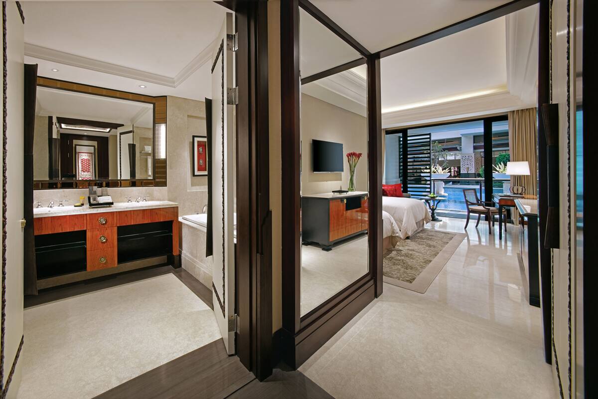 A modern hotel room with a large mirror, a double sink vanity, a bed, a desk, and a view of a pool.