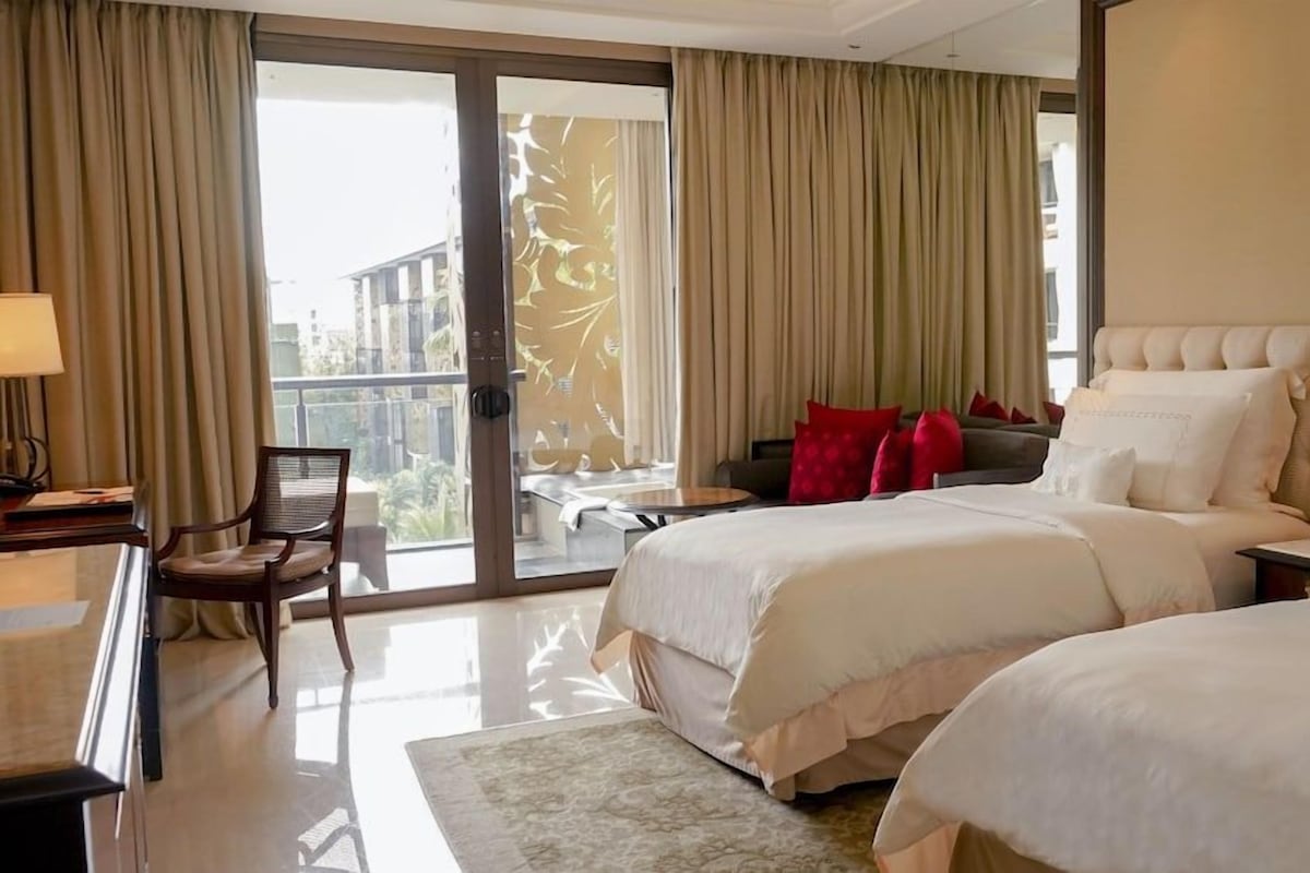 A hotel room with two beds, a desk, a chair, a balcony with curtains, and a view of buildings outside.