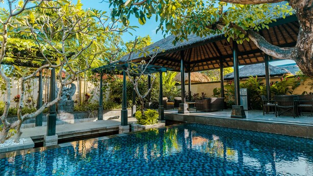 Una piscina con borde de azulejos, rodeada de vegetación y un área de estar cubierta con sillas y mesas.