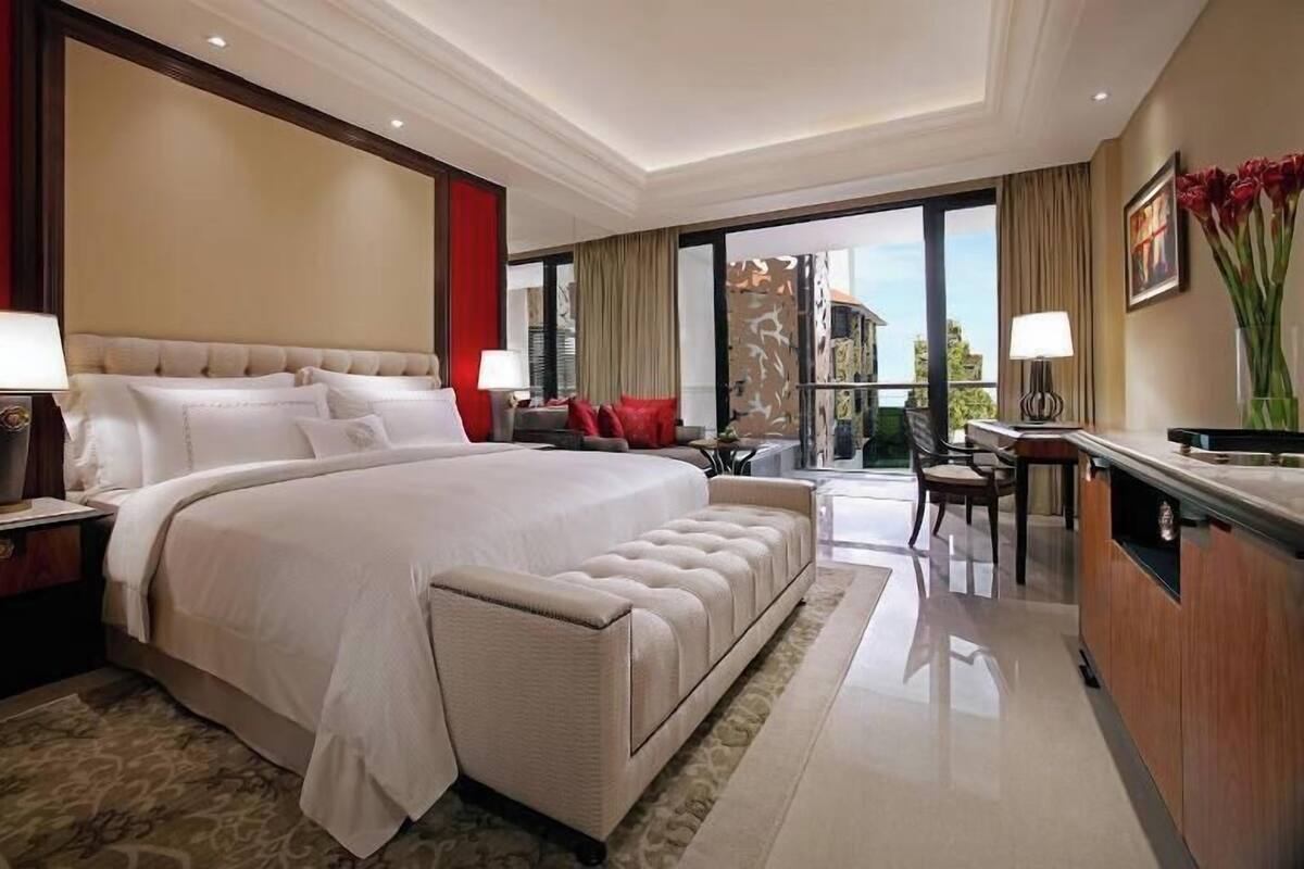 A spacious hotel room with a large bed, a sitting area, a dining table, and a view of the outdoors.