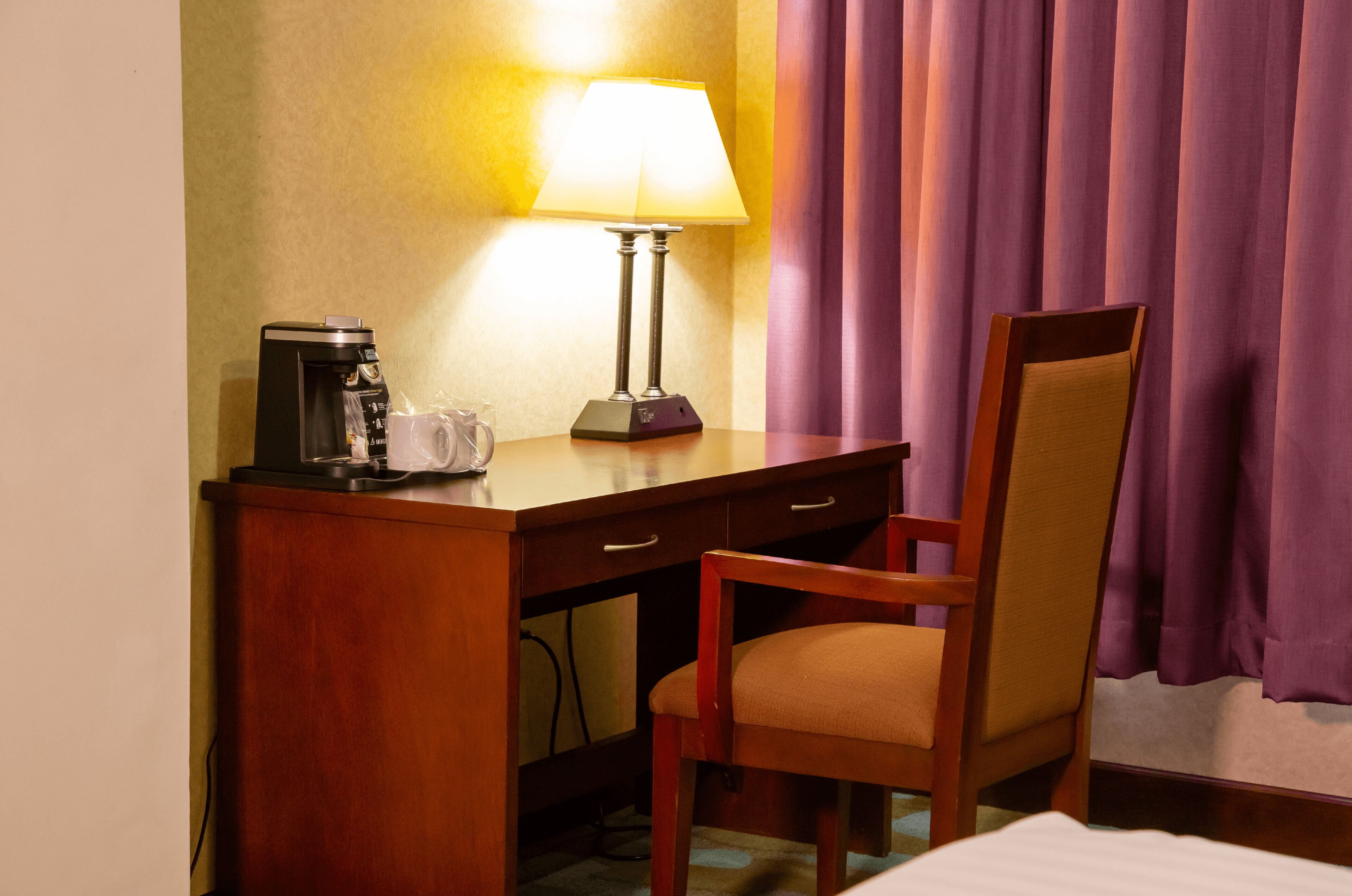 A hotel room with a wooden desk, a lamp, a chair, a bed, and purple curtains.