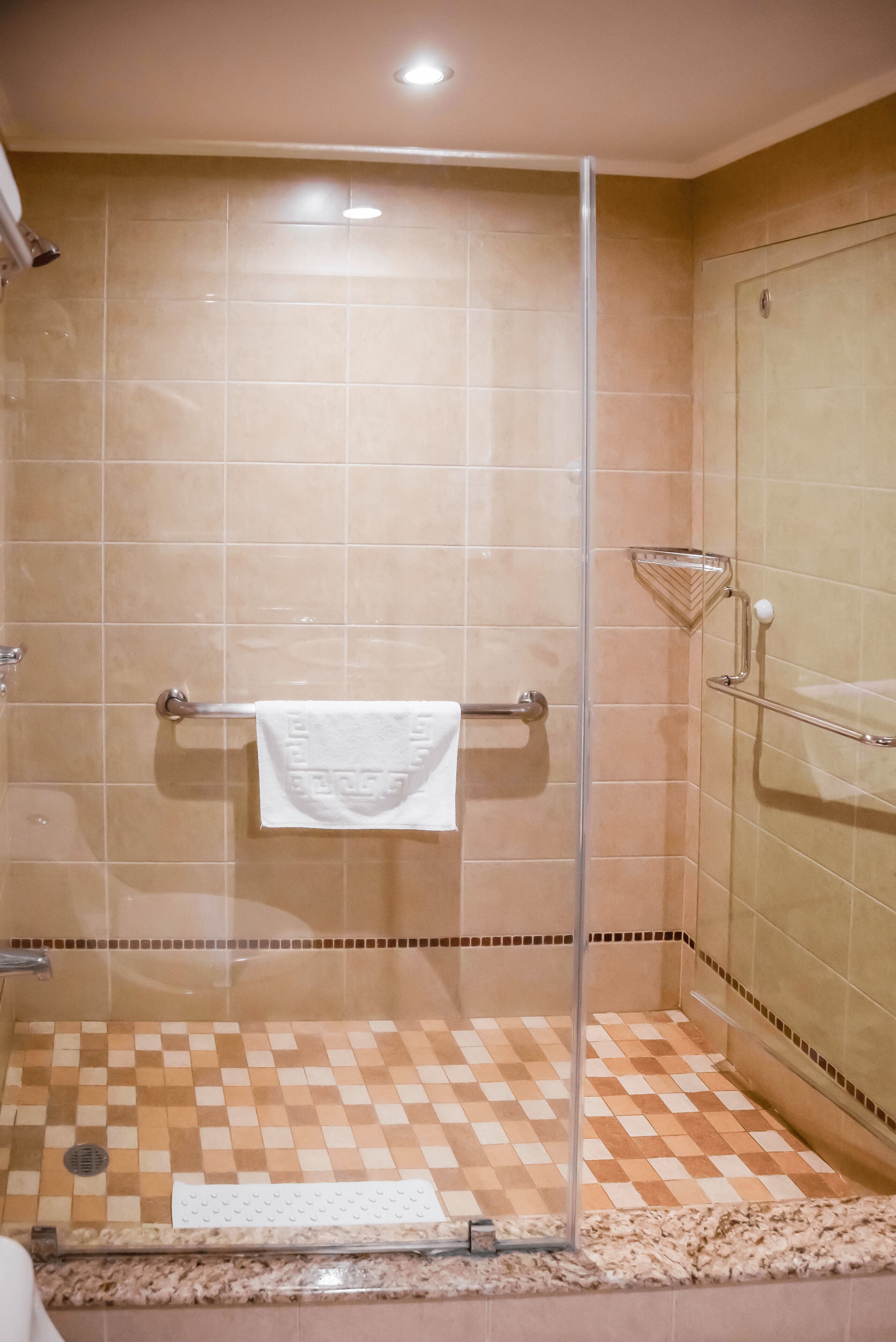 Una ducha con cerramiento de vidrio, azulejos color beige, una toalla blanca en un perchero y un suelo con diseño a cuadros.