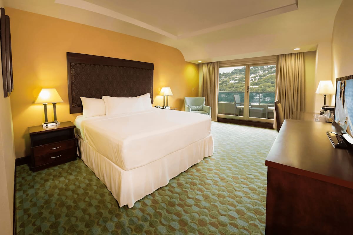 A hotel room with a large bed, two bedside tables with lamps, a chair, a desk with a TV, and a balcony with a view.