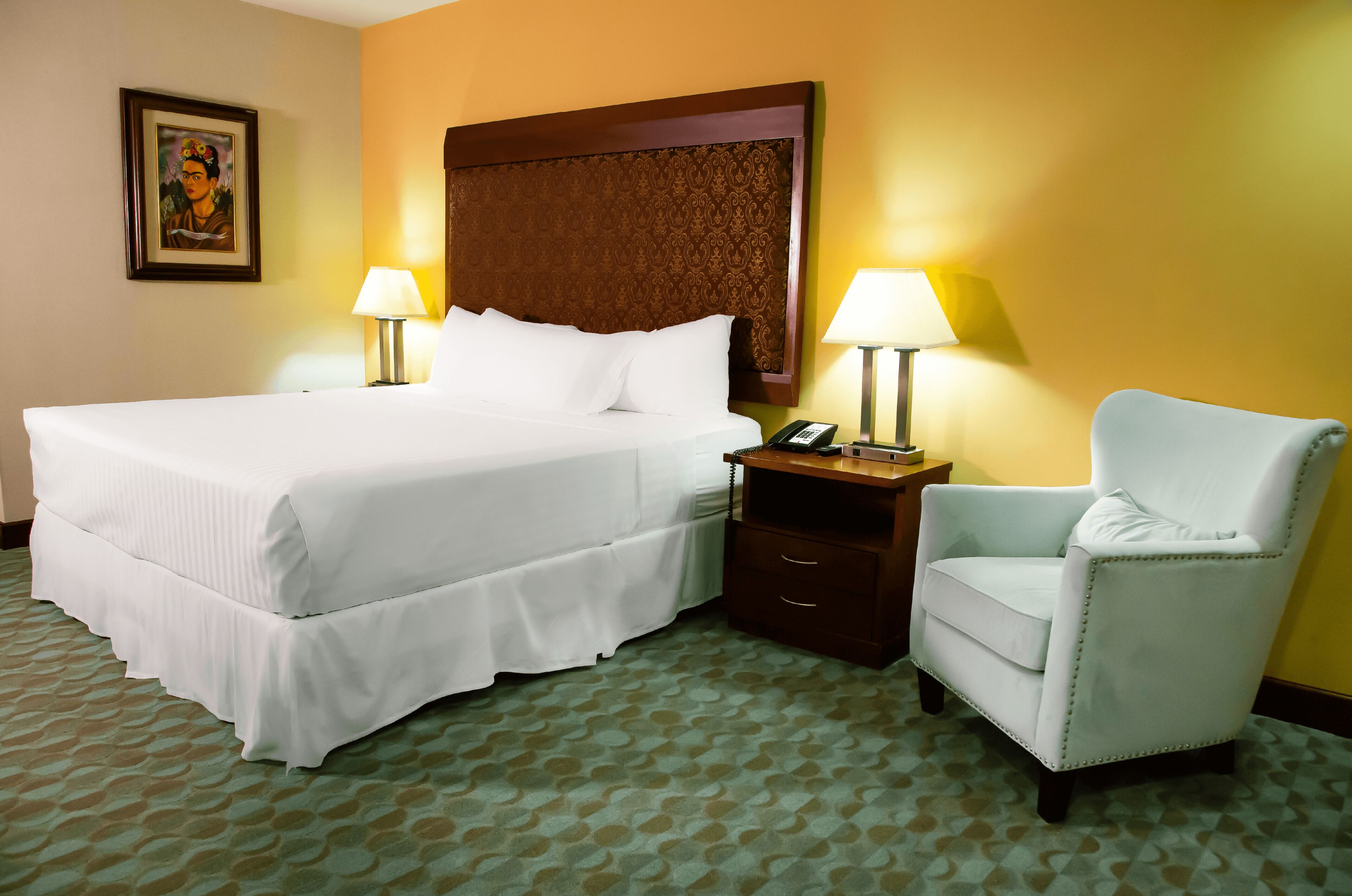 Una habitación de hotel con una cama grande, dos lámparas de noche, una mesita de noche de madera, un sillón blanco y una alfombra estampada.