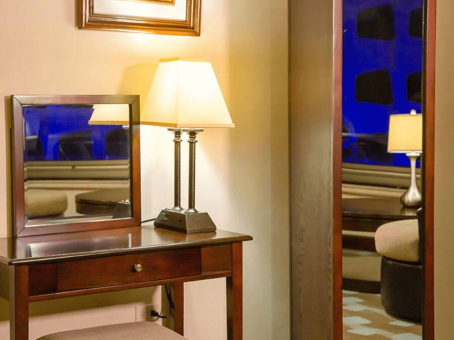 A hotel room with a desk, chair, lamp, and a mirror.