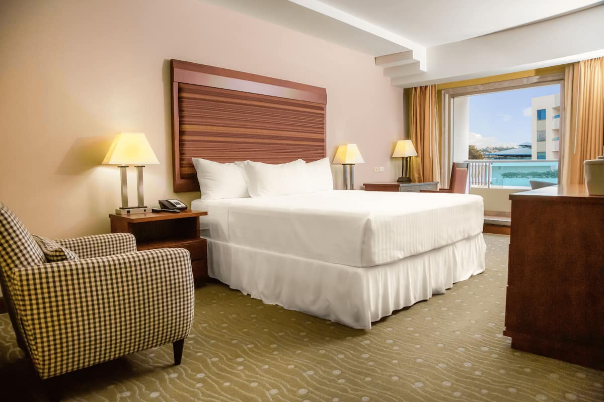 A hotel room with a large bed, two bedside tables with lamps, a checkered armchair, and a view of the sea through a balcony.
