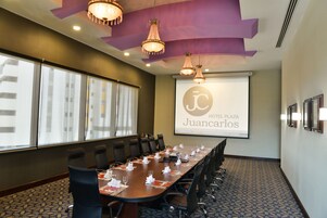 Uma sala de conferências com uma mesa comprida, cadeiras e uma tela de projetor exibindo 'Hotel Plaza Juancarlos'.