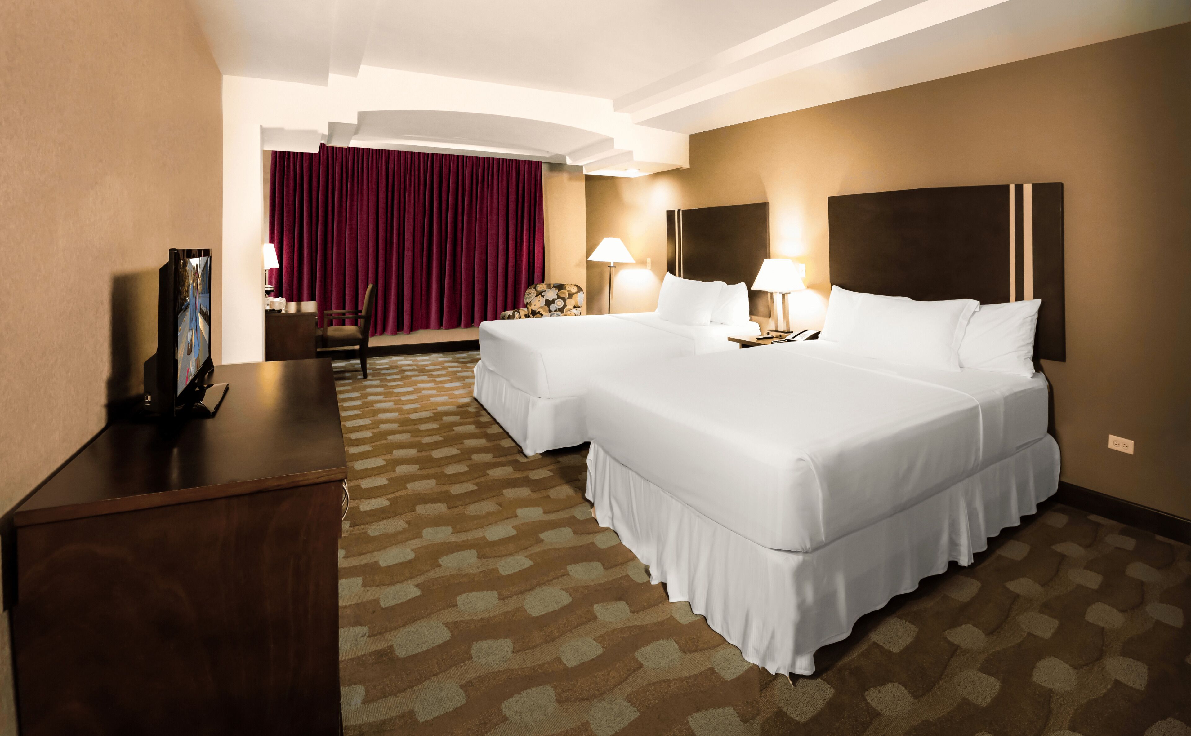 A hotel room with two beds, a desk, a TV, and a patterned carpet.
