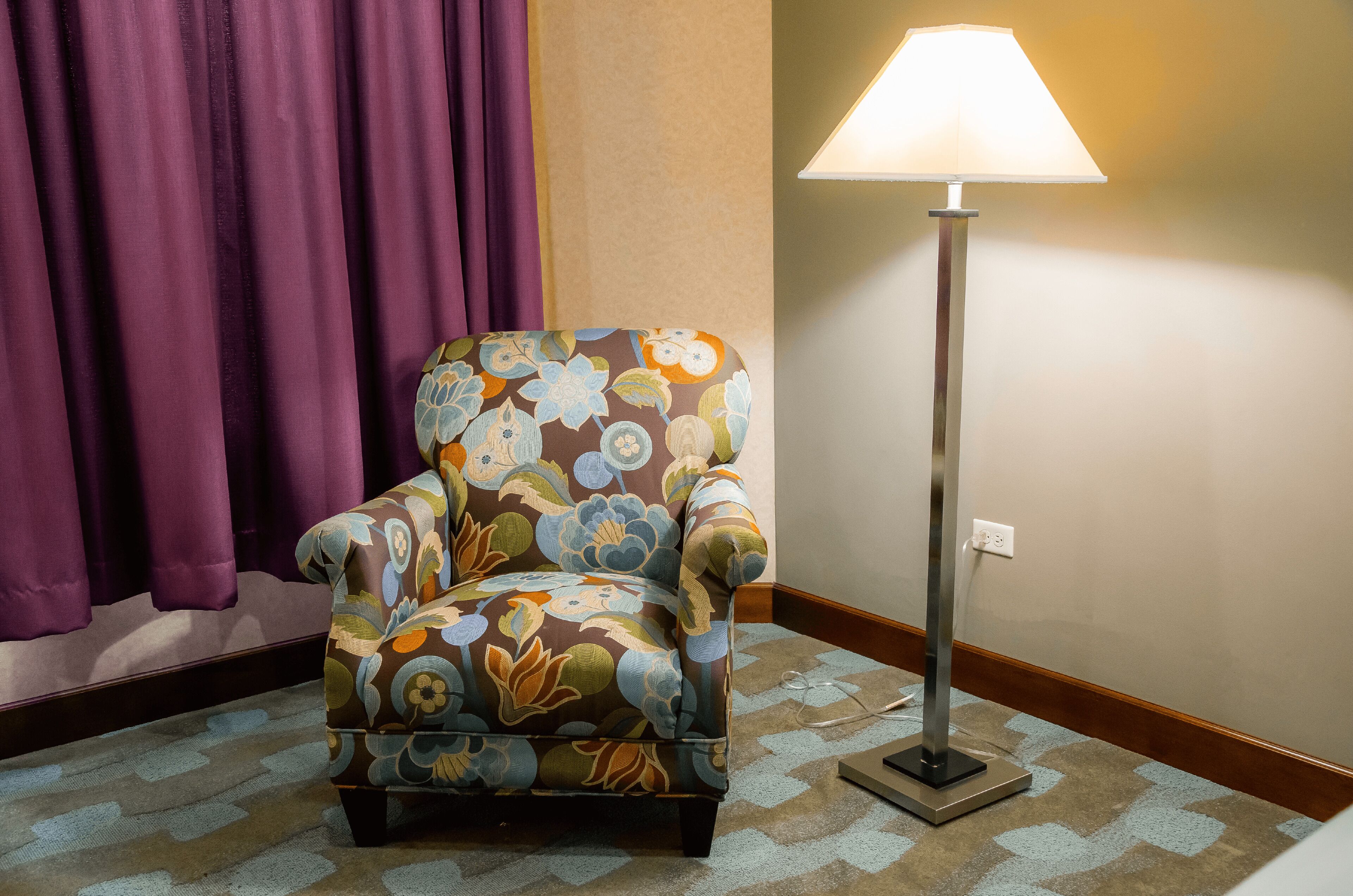 A floral armchair with a patterned cushion, a standing floor lamp, and a patterned carpet in a room with purple curtains and a beige wall.