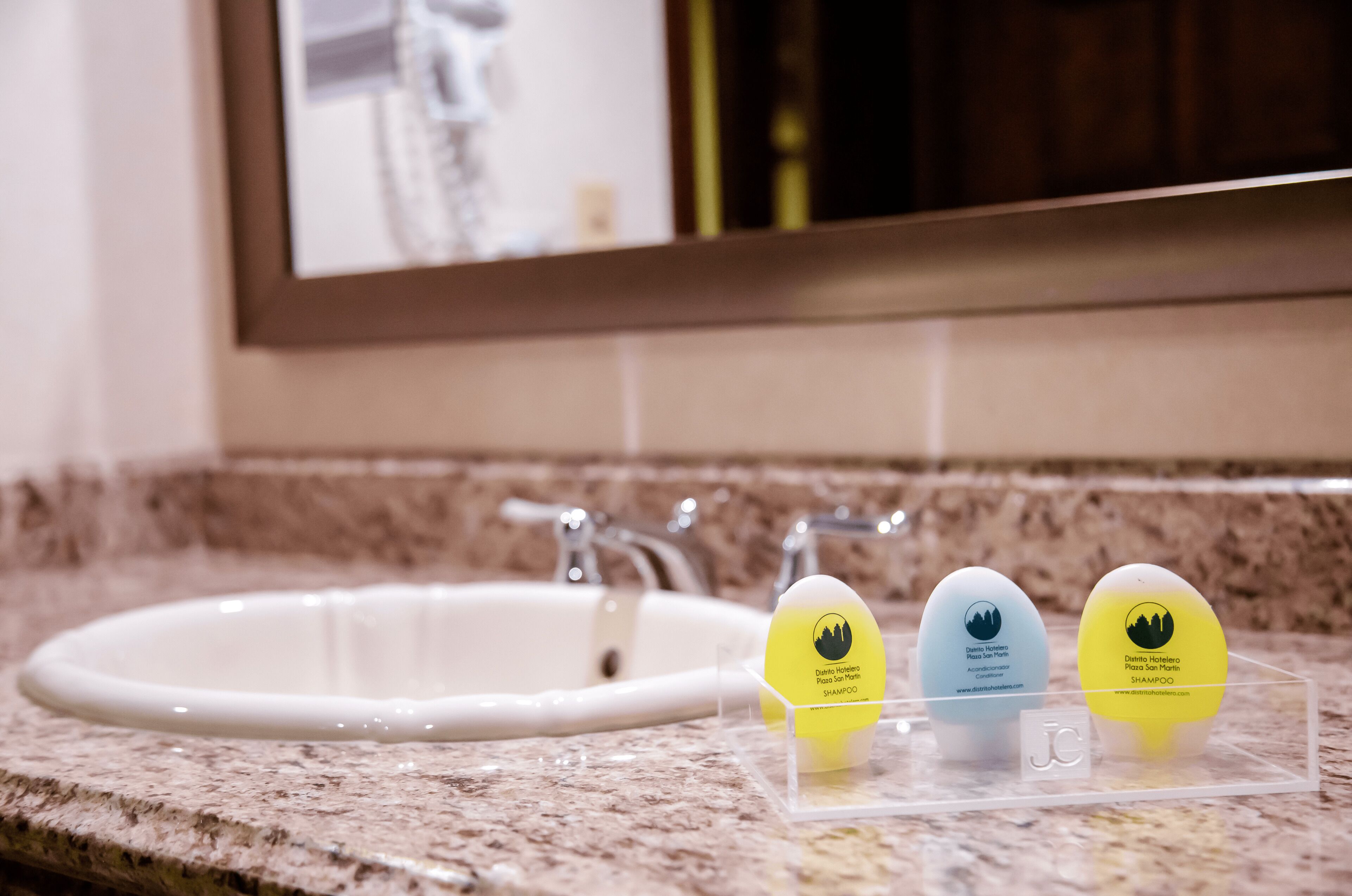 A bathroom sink with a mirror above it. There are three egg-shaped containers on a clear tray, each labeled with a hotel name and website.