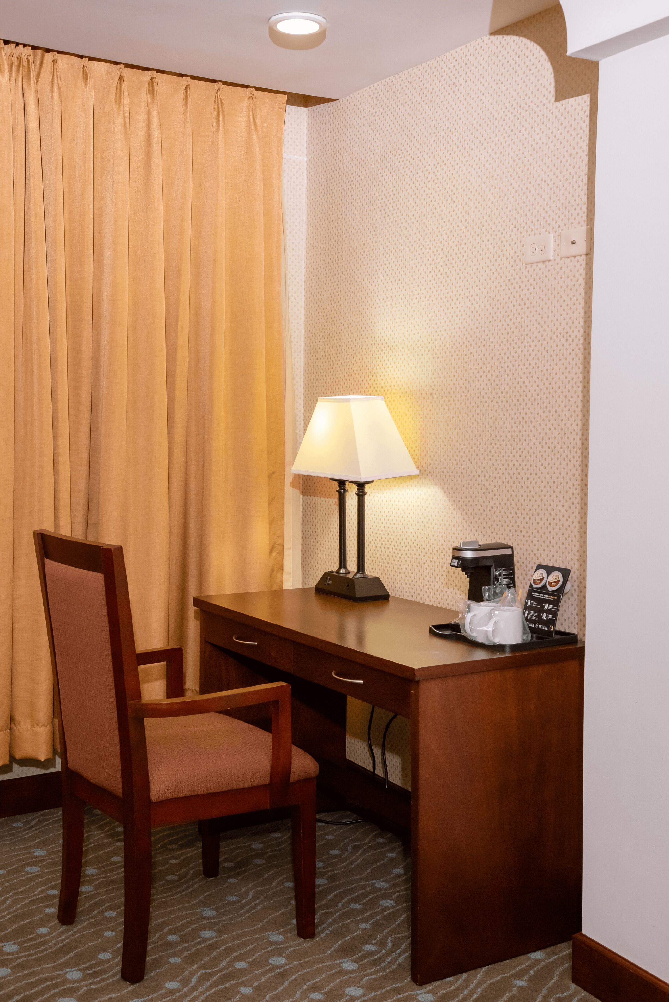 A hotel room with a wooden desk, a chair, a lamp, and a coffee machine.