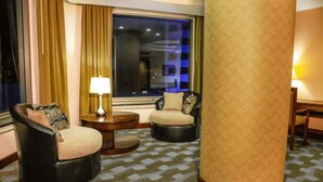 Habitación de hotel con un ventanal grande, una mesa redonda con lámpara, dos sillones y una alfombra estampada.