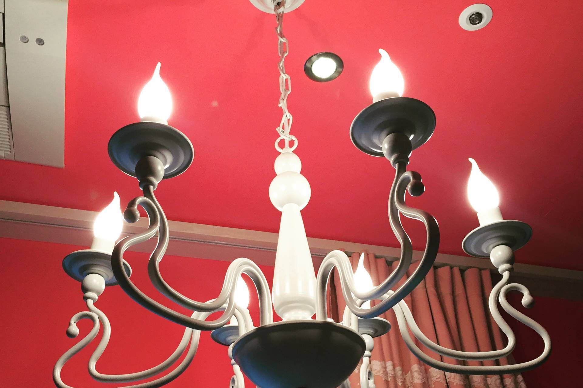 Een kroonluchter met meerdere lichtjes, een rode muur en een zichtbare ventilatieopening in het plafond.
