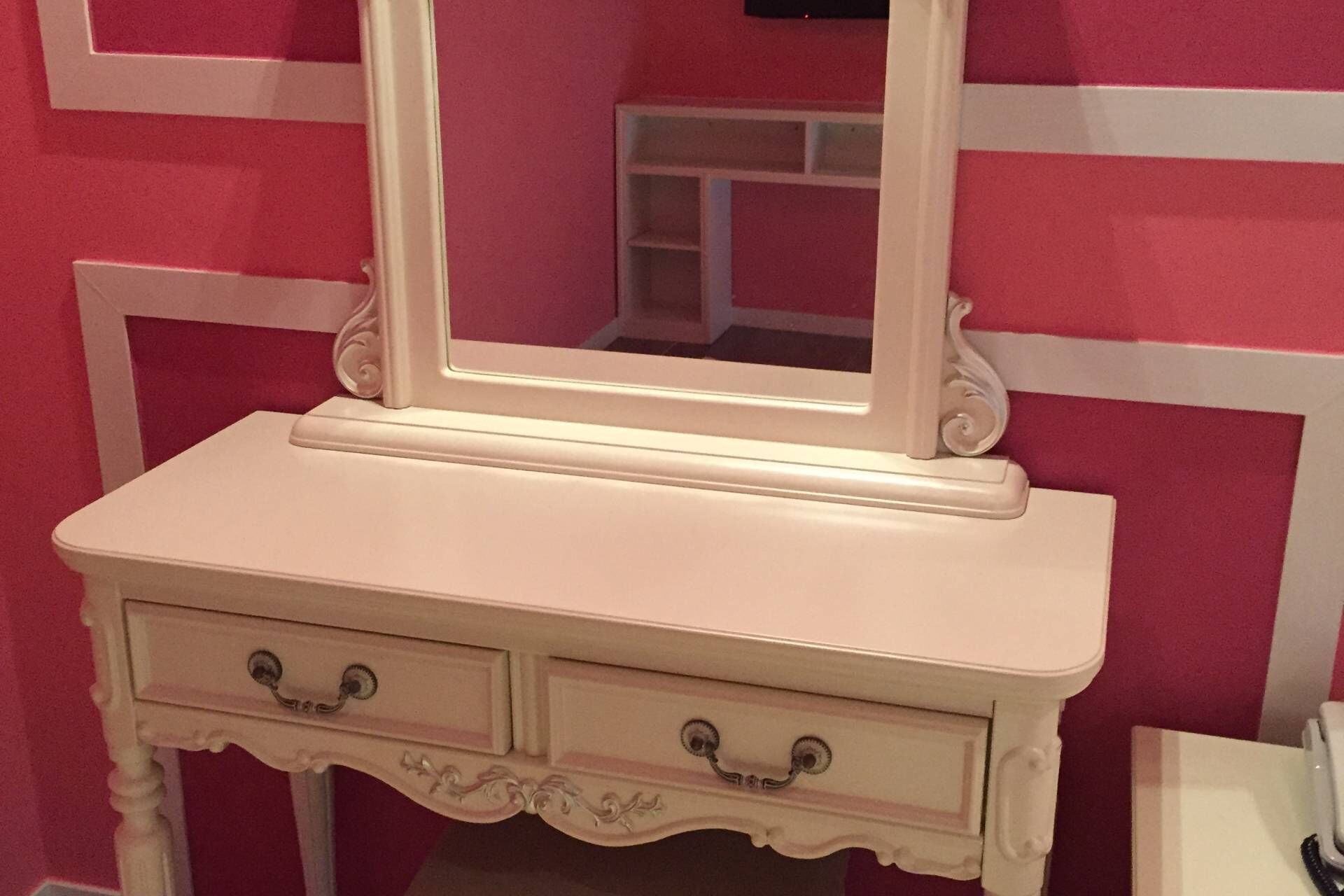 Une coiffeuse blanche, dotée d’un miroir et de deux tiroirs, est placée contre un mur rose orné de moulures blanches.
