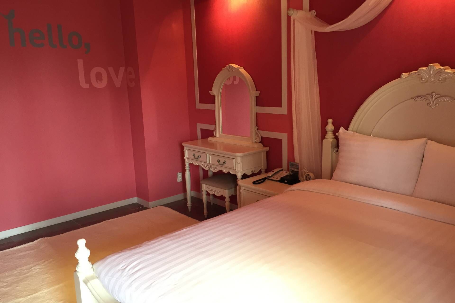 A bedroom with a bed, a vanity with a mirror, a nightstand, and a wall with the text 'hello, love'.