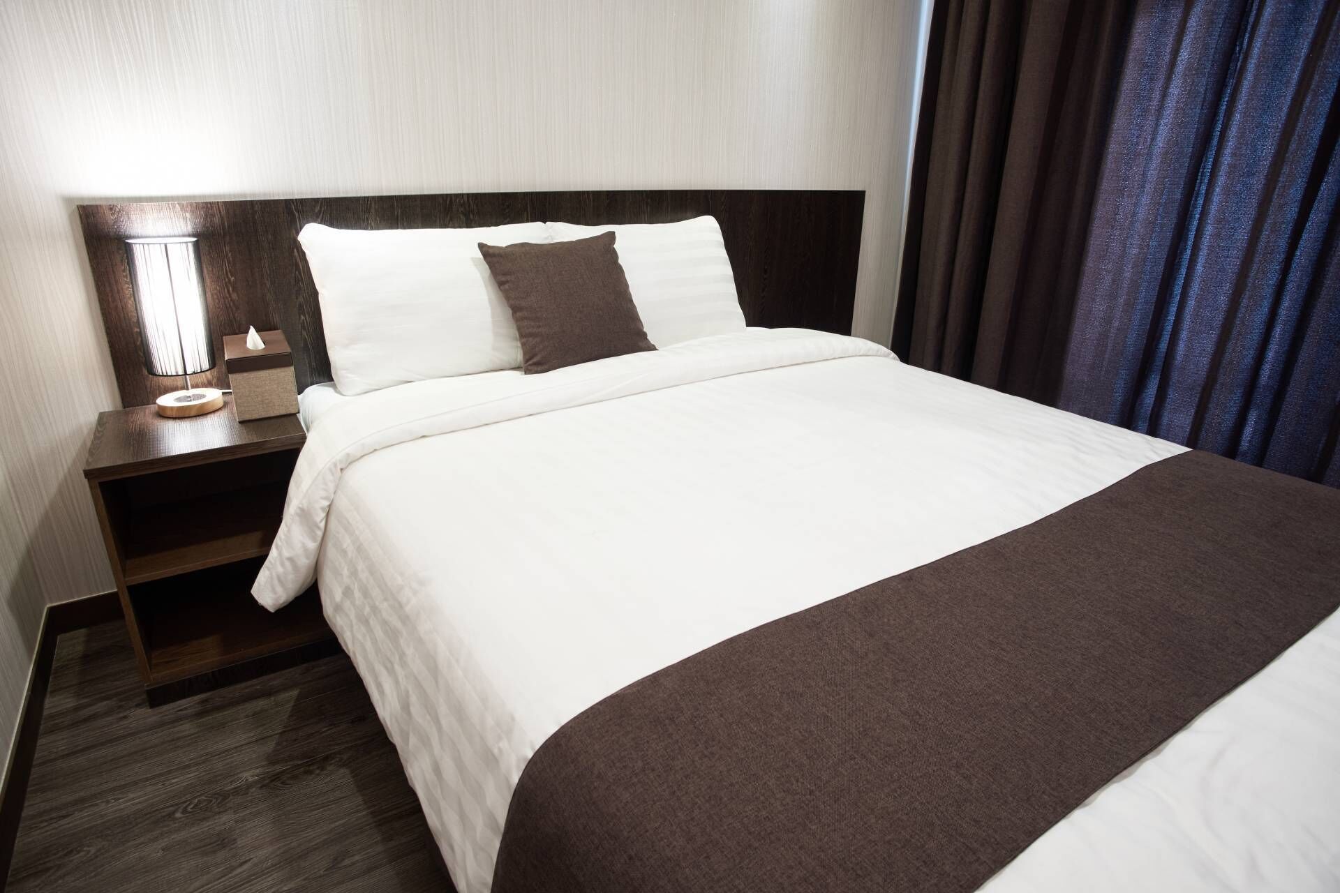 A neatly made bed with white and brown bedding, a bedside lamp, and a wooden nightstand.