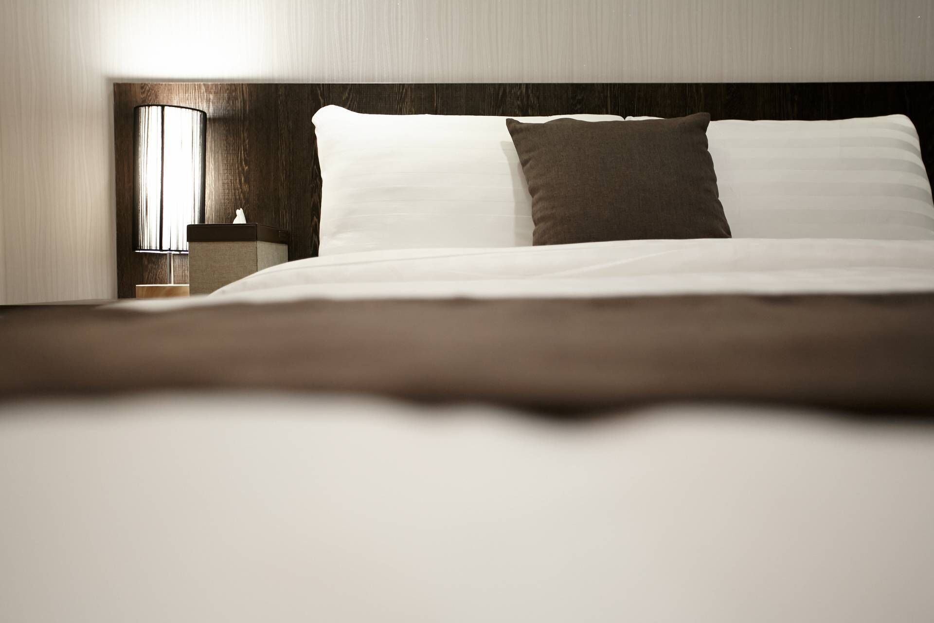 A neatly made bed with white and brown pillows, a wooden headboard, and a bedside table with a lamp.