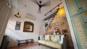 Una sala de estar con sofá, banqueta, alfombra estampada, ventilador de techo, espejo montado en la pared y pared de ladrillo.