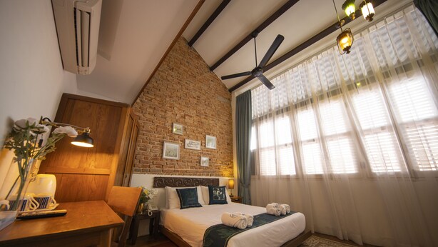 Un dormitorio con pared de ladrillo, cabecera de madera, cama con sábanas blancas, escritorio con lámpara y ventana con cortinas traslúcidas.
