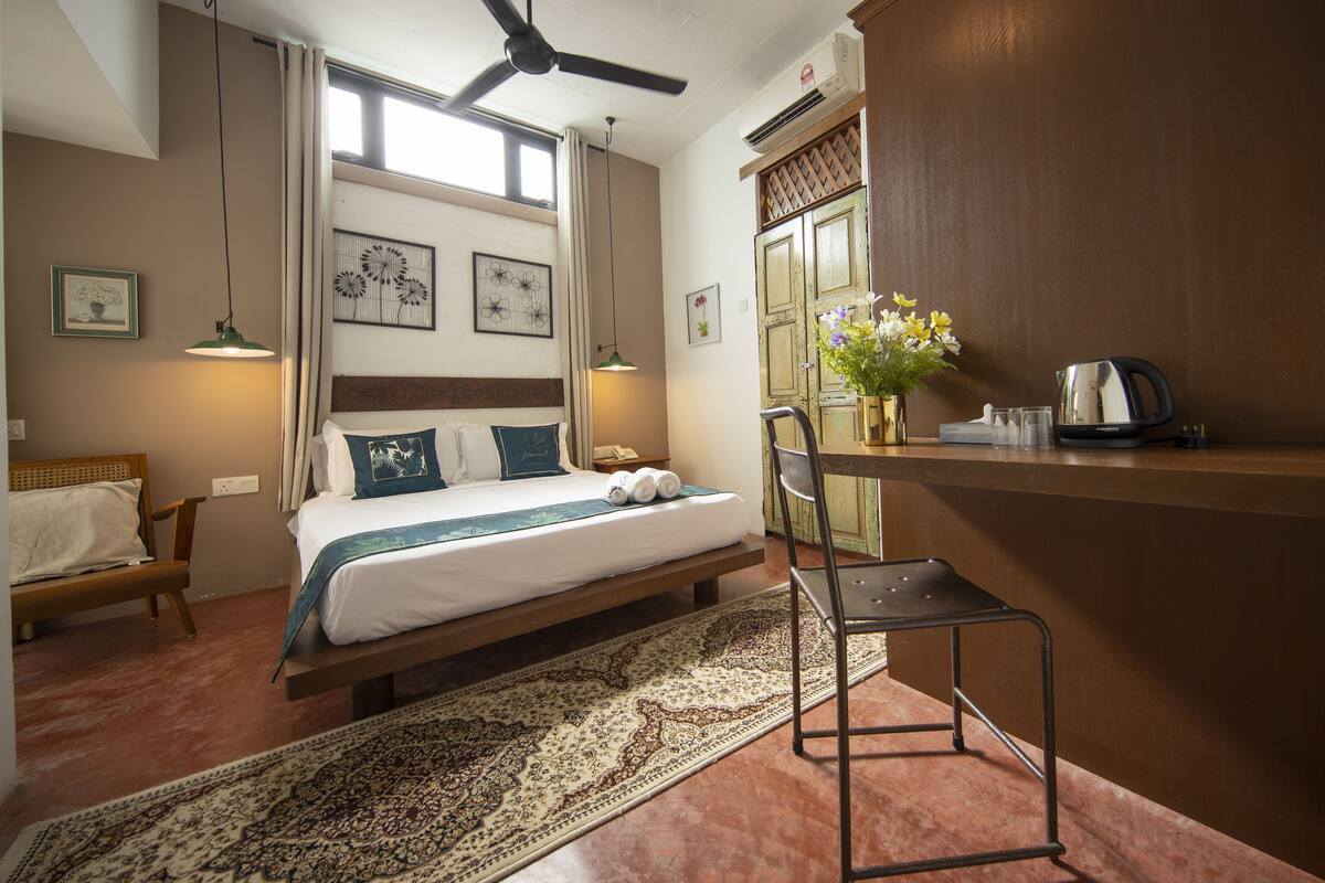 Une chambre d’hôtel comprenant un lit, un bureau, une chaise, une lampe de bureau, un ventilateur de plafond, une fenêtre et un climatiseur fixé au mur.