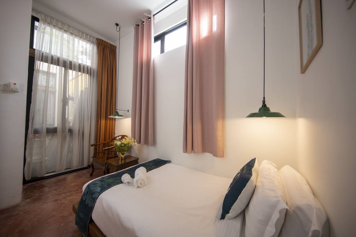 Une chambre d’hôtel avec un lit, un bureau, une chaise, une lampe et une fenêtre avec des rideaux.