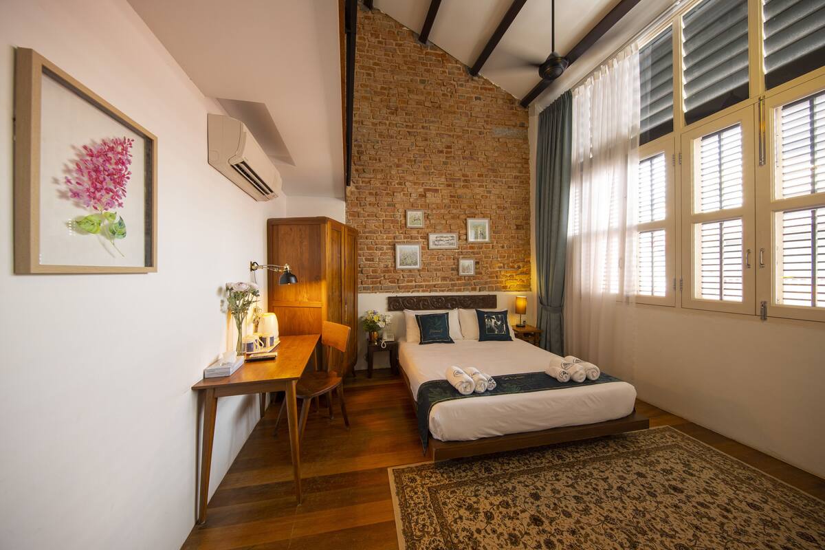 Une chambre avec un mur en briques, un bureau en bois, un lit avec des oreillers et une fenêtre avec des stores.