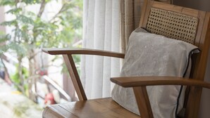 Una silla de madera con respaldo trenzado y cojín, ubicada cerca de una ventana con cortinas traslúcidas, que ofrece vistas a follaje exterior.