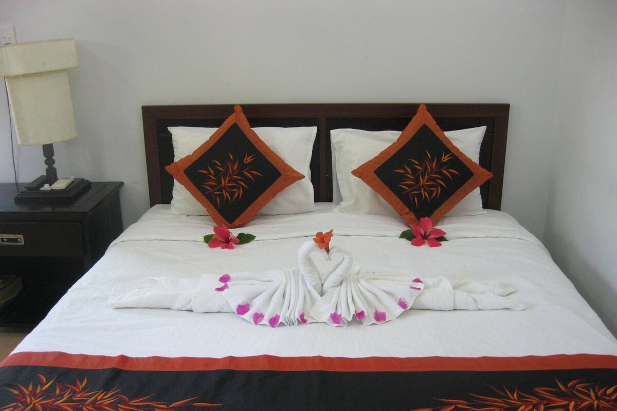 A neatly made bed with a towel arranged in a swan shape, two decorative pillows, and a nightstand with a lamp.
