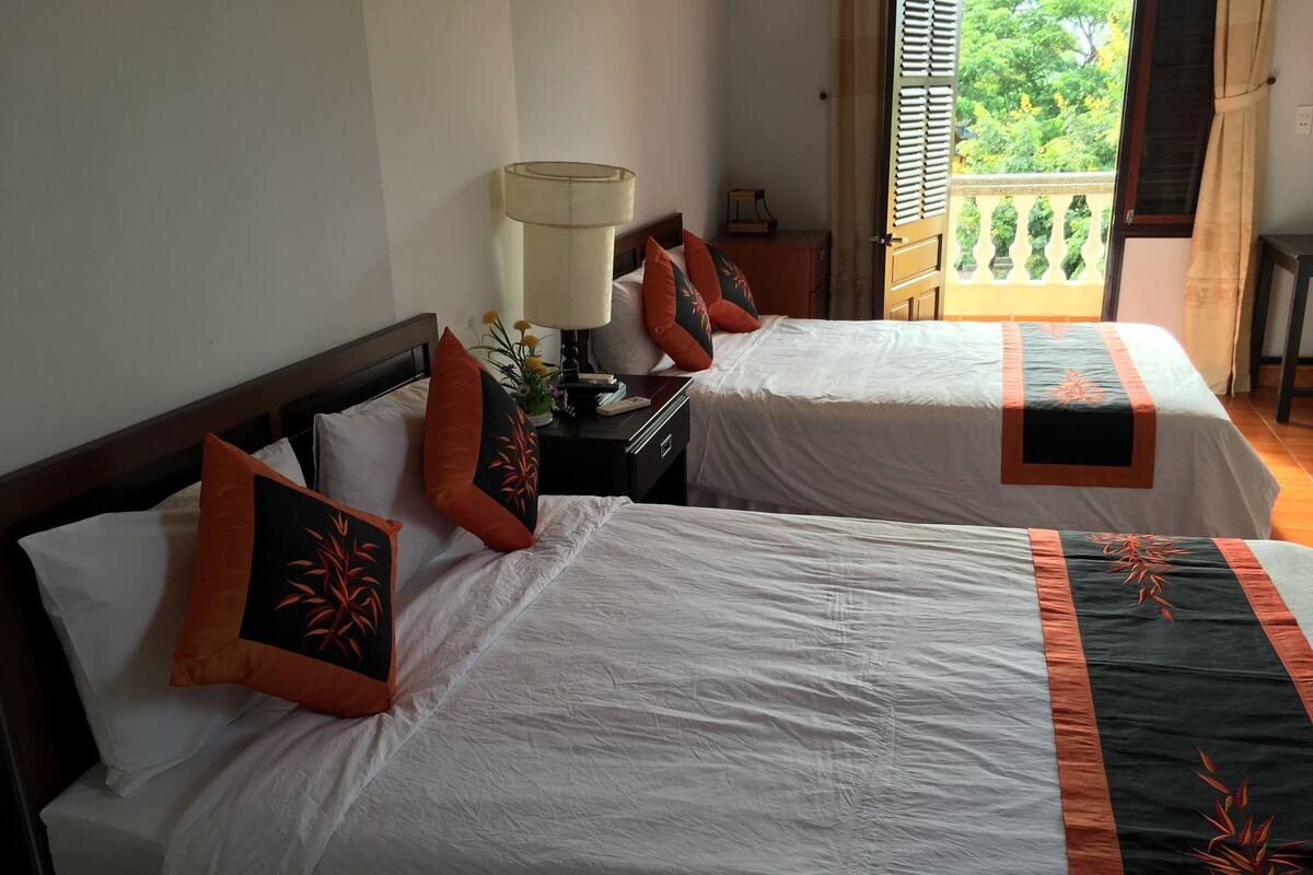 A hotel room with two beds, each with white linens and orange decorative pillows. There is a nightstand with a lamp and a small table with a phone and a decorative item. A window with a view of greenery is visible.