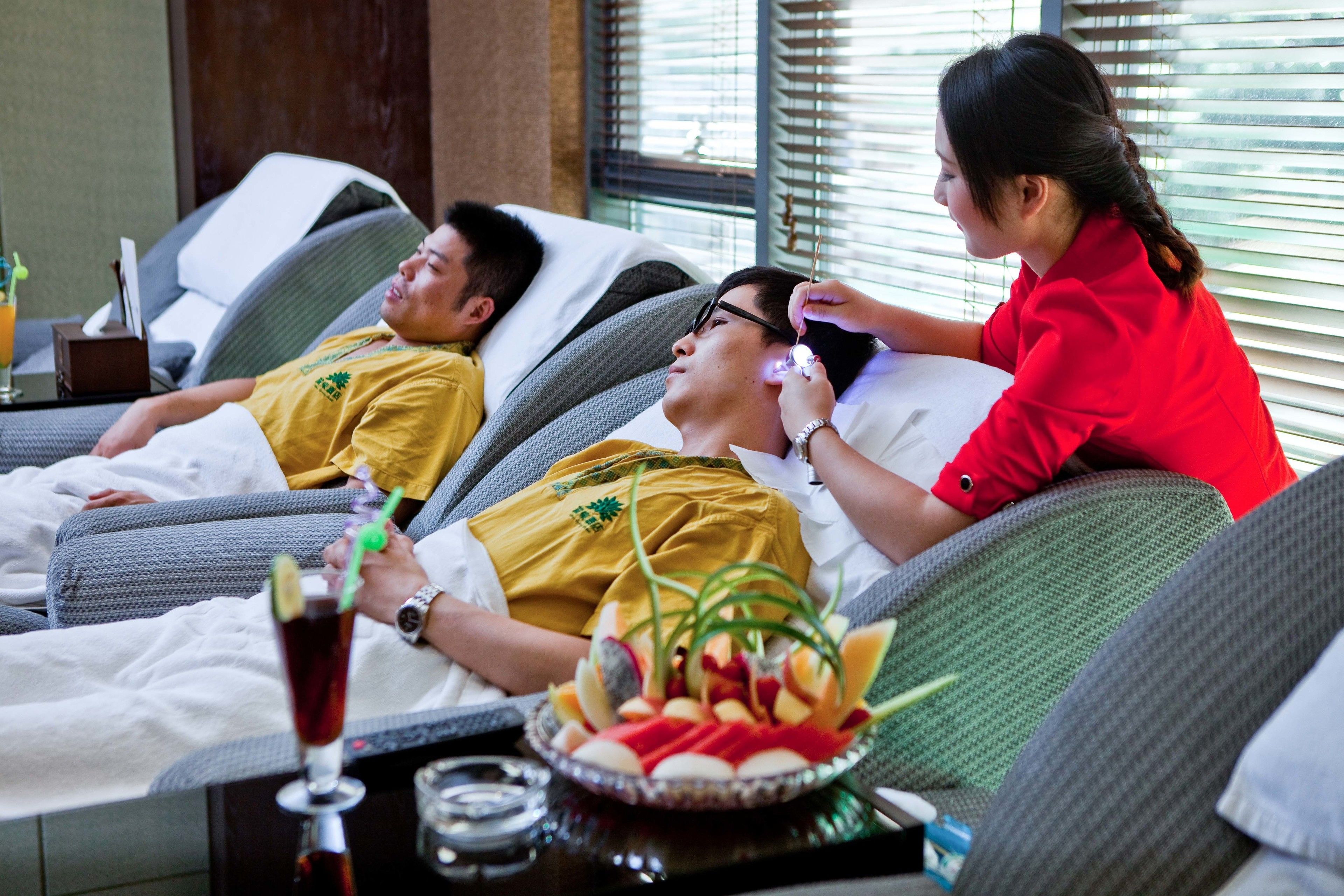 Een massageruimte met twee cliënten die op verstelbare stoelen liggen, een massager geeft één cliënt een massage, en een dienblad met een drankje en fruit op de tafel.