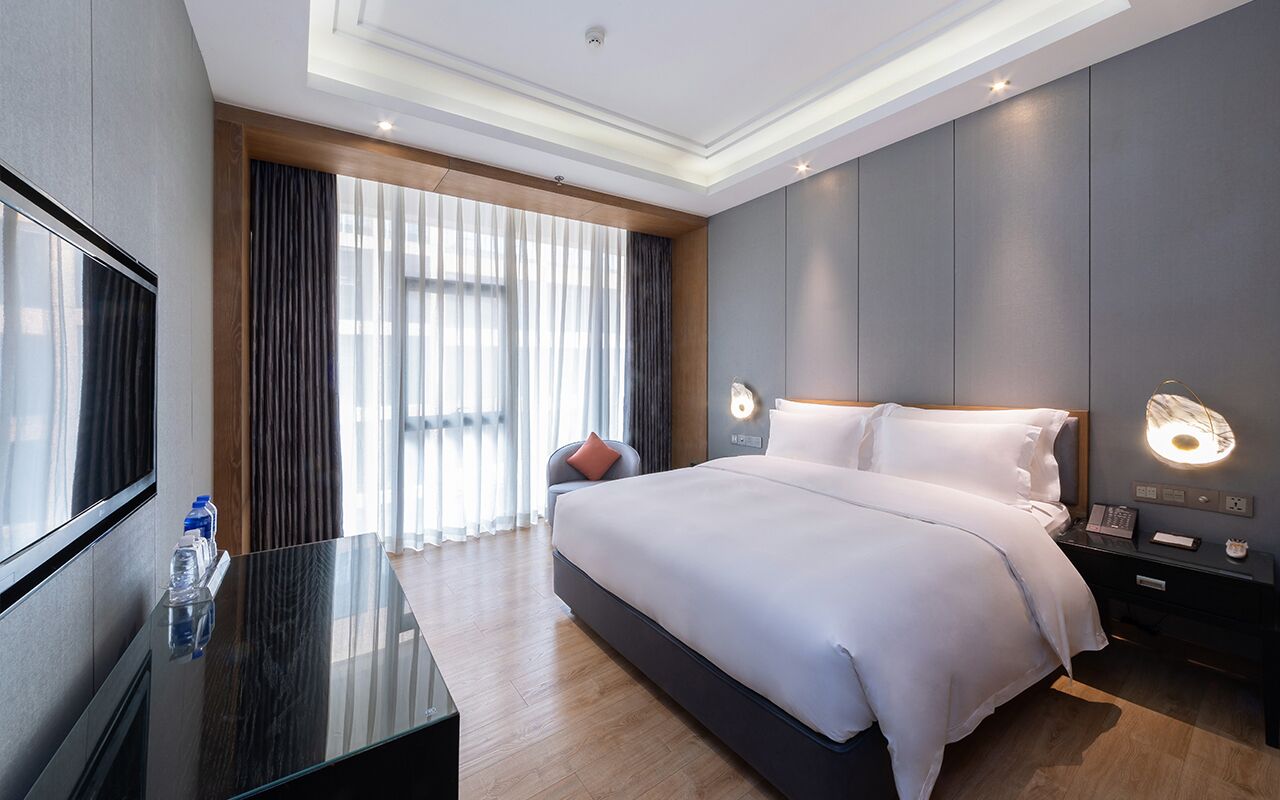 A modern hotel room with a large bed, a flat-screen TV, a bedside table, and a window with curtains.