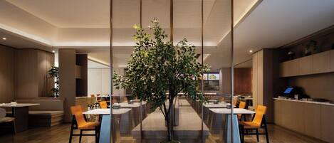 A modern dining area with a central table surrounded by chairs, a large potted tree, and a partition with a mesh design.