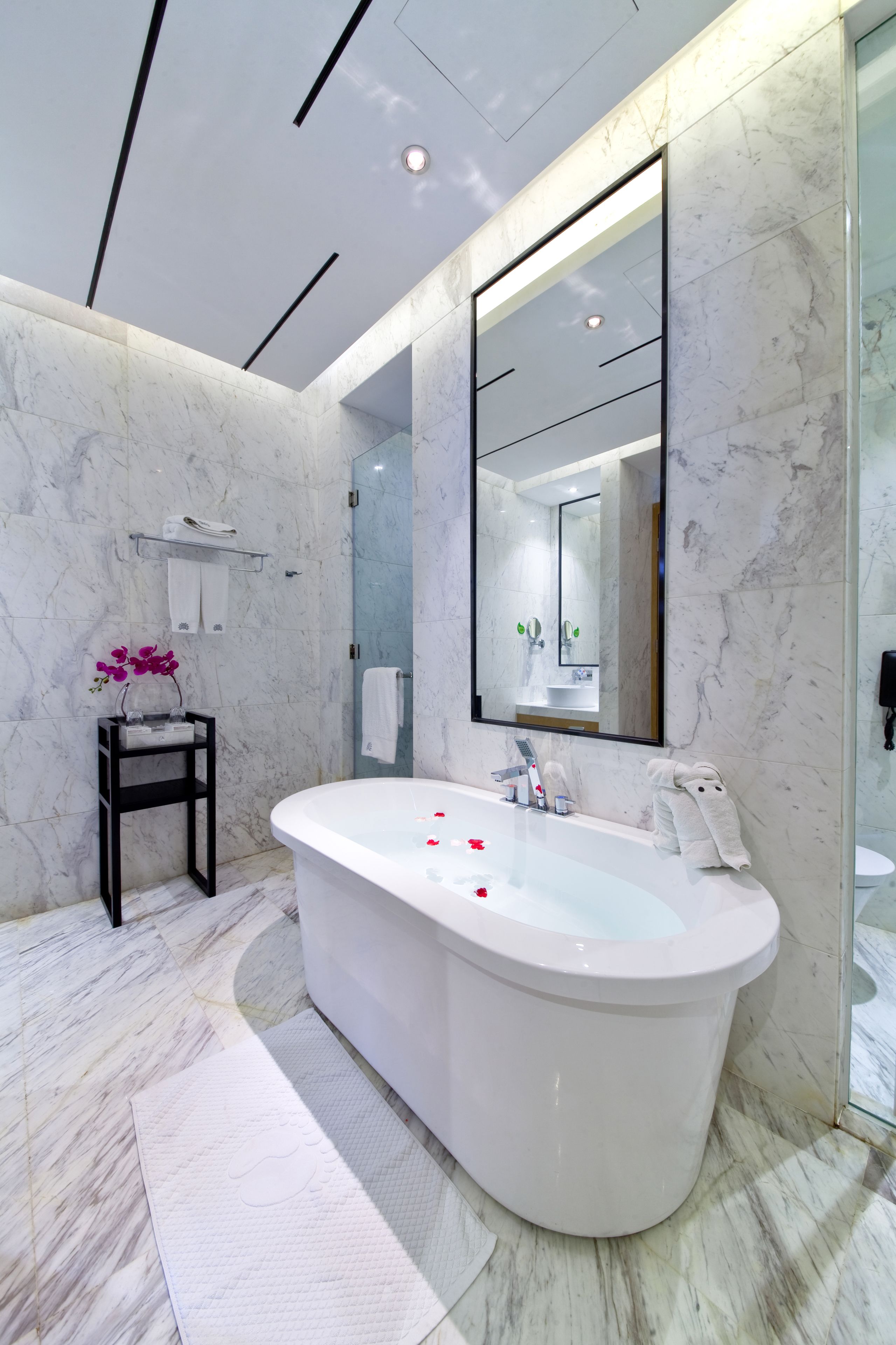 A modern bathroom with a freestanding bathtub, marble walls, a large mirror, and a small shelf with towels and an orchid.