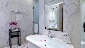 A modern bathroom with a freestanding bathtub, marble walls, a large mirror, and a small shelf with towels and an orchid.