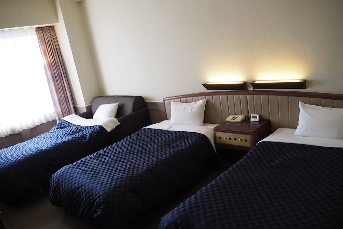 Ett hotellrum med två sängar, en stol, ett nattduksbord med en telefon och ett fönster med gardiner.