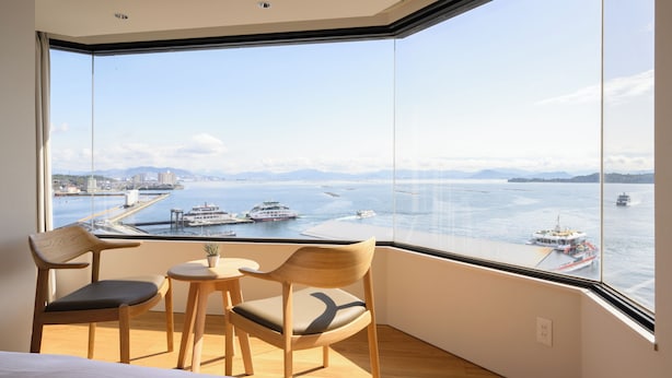 A room with a large window overlooking a body of water with boats, a view of a distant city, and a small table with chairs.