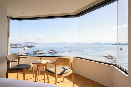 A room with a large window overlooking a body of water with boats, a view of a distant city, and a small table with chairs.