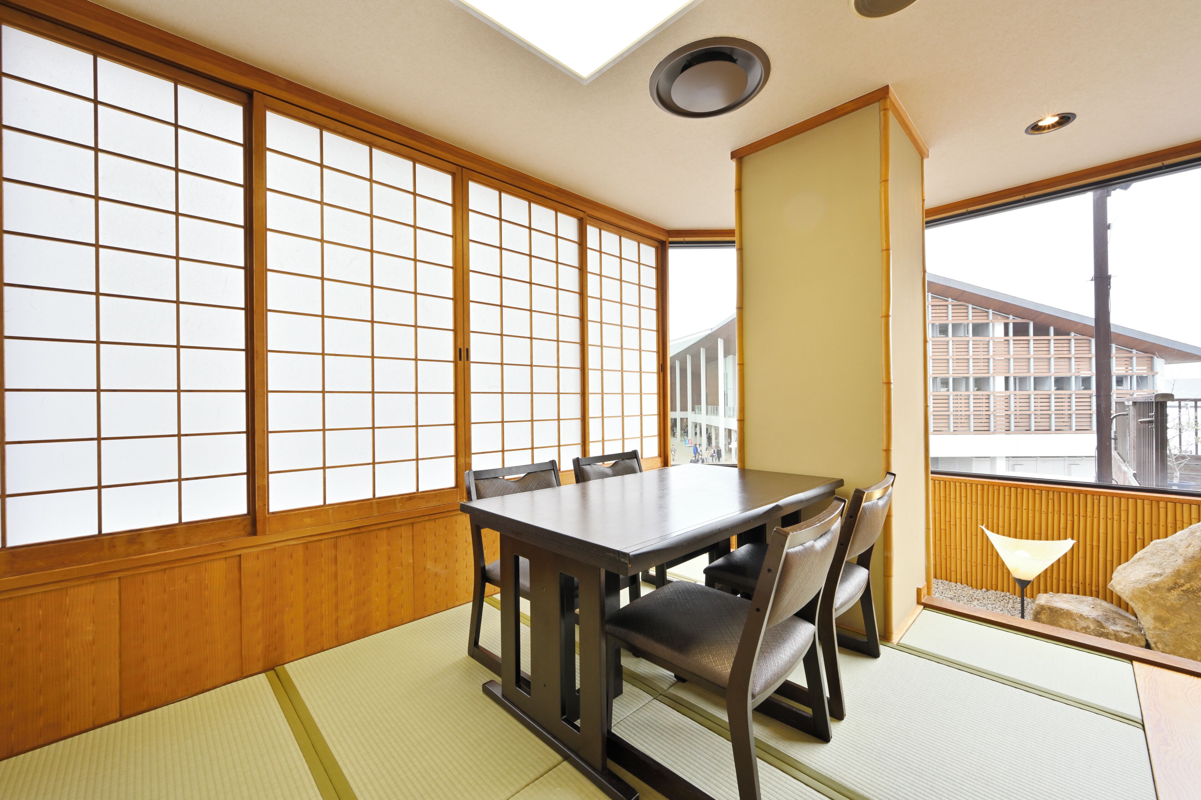 A traditional Japanese room with tatami flooring, a low table with chairs, sliding shoji doors, and a view of a building outside.