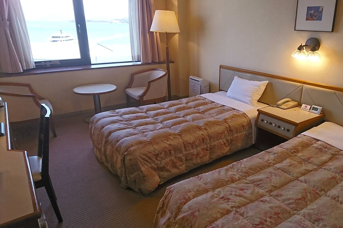 Ett hotellrum med två sängar, ett skrivbord, en stol, en lampa och utsikt över havet.